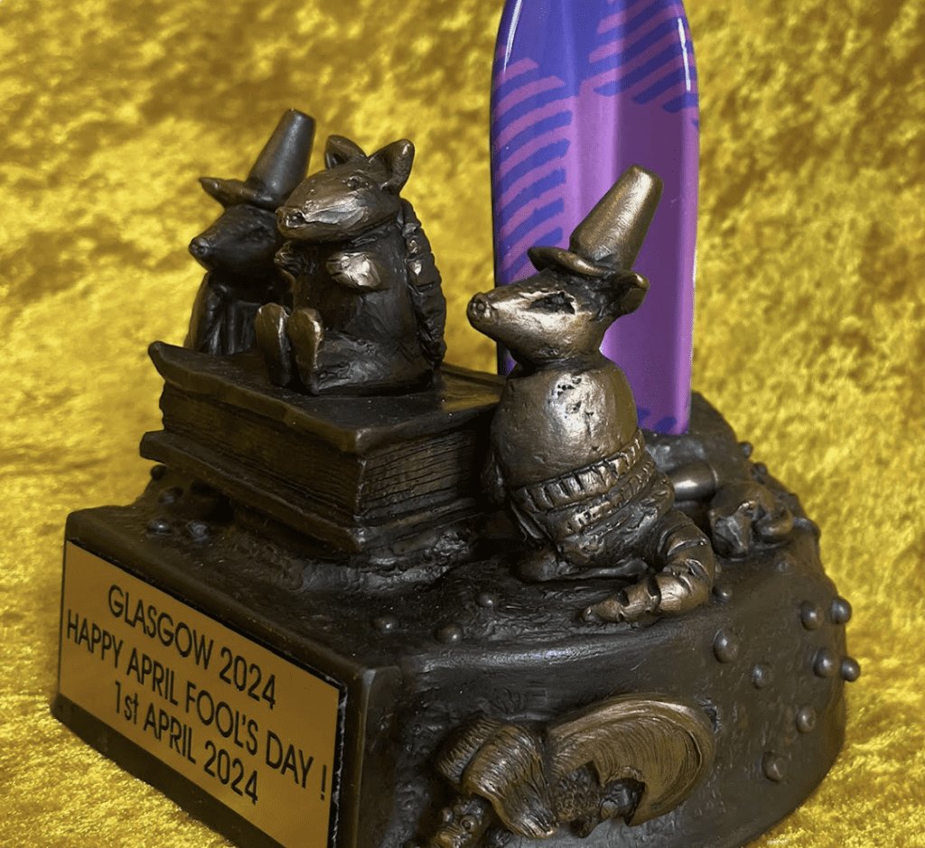 A angled view of an ornamental trophy celebrating April Fool's Day in Glasgow for the year 2024. The trophy features the bottom half of a, vertically standing, patterned rocket in hues of purple and blue with tartan pattern. The base is a dark, metallic color, depicting three armadillos, two in traffic cone hats flank a centered armadillo sitting atop a book and a depiction of a flying haggis on the side. Center, a plaque reads 'GLASGOW 2024 HAPPY APRIL FOOLS' DAY! 1st APRIL 2024' against a textured gold background.
